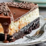 A decadent salted caramel chocolate cheesecake topped with drizzled caramel and chocolate shavings on a white plate.