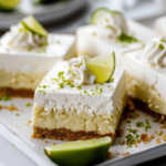 A close-up of key lime pie bars topped with whipped cream and lime zest on a white plate.
